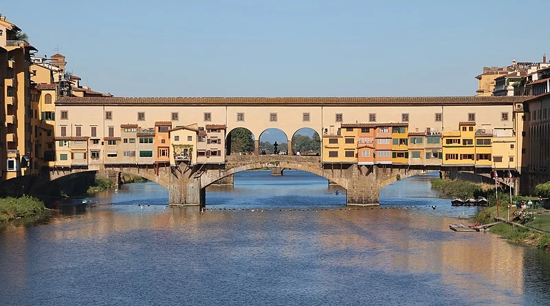 A historic medieval stone bridge spanning the Arno River in Florence, Italy, lined with charming shops and featuring large central arches.