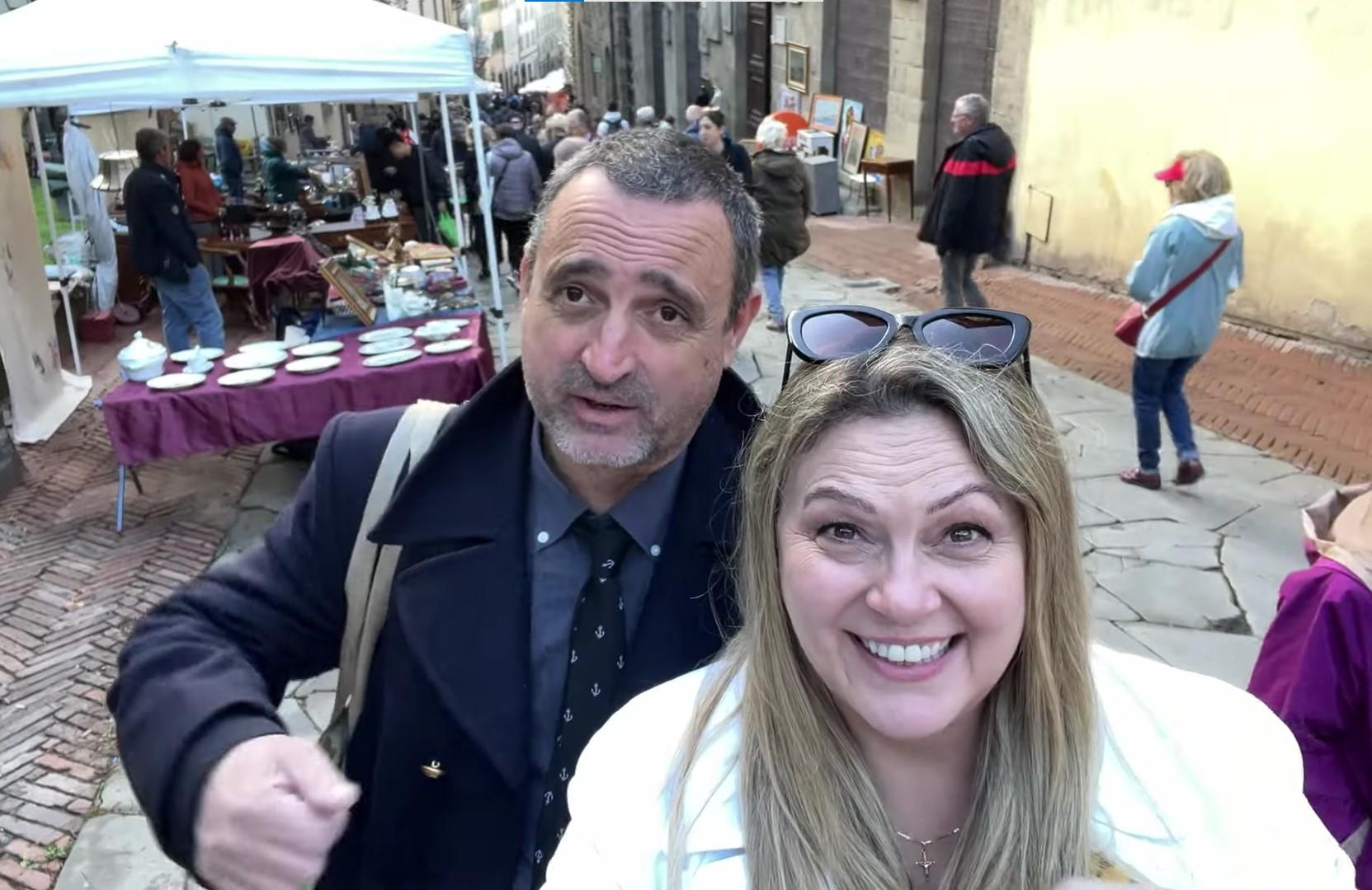 A man and a woman take a selfie at an outdoor antique fair in Arezzo, Italy. The woman in the foreground is smiling, wearing a white coat with sunglasses on her head, while the man beside her wears a dark coat and gestures as if speaking.