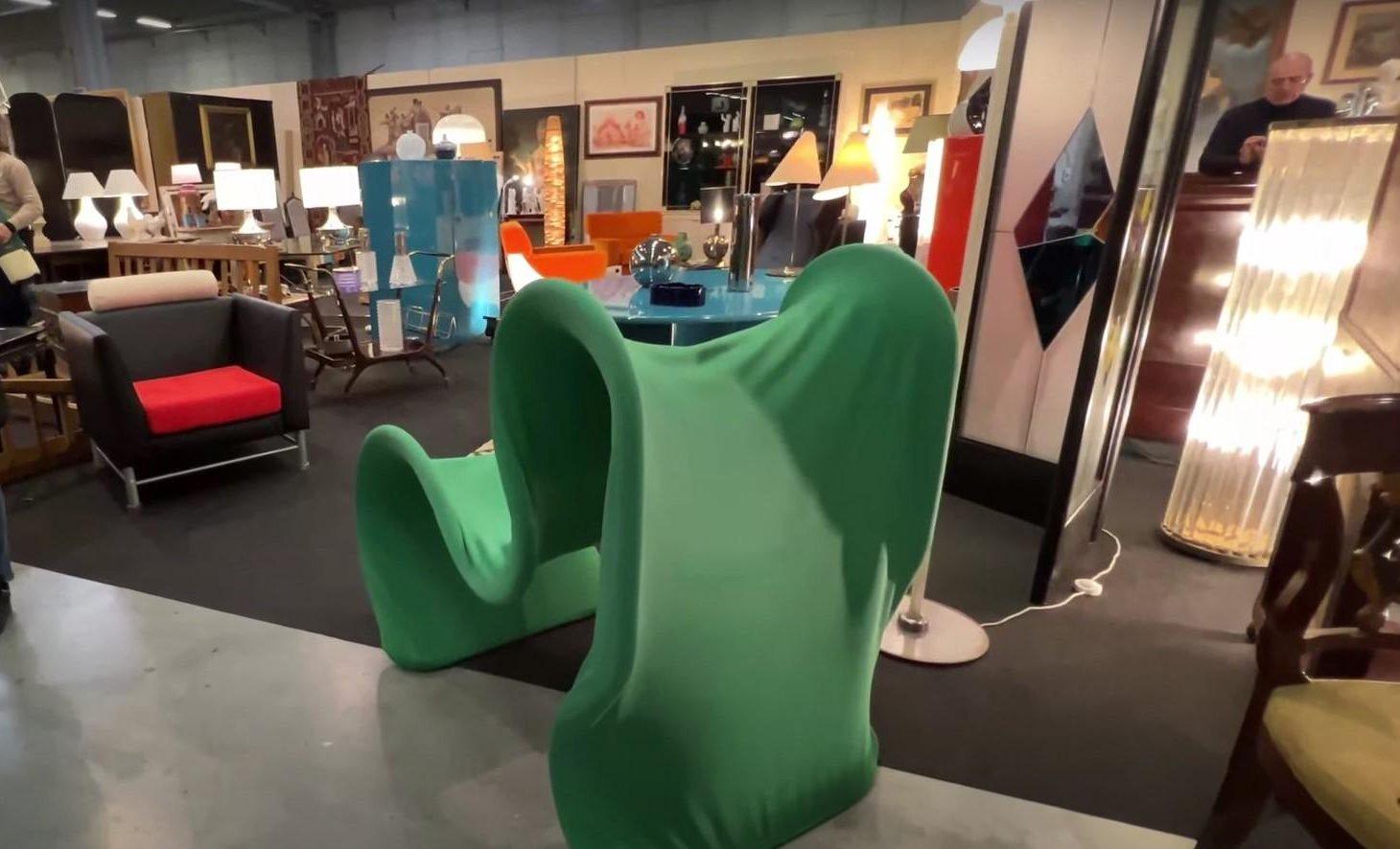 A vintage furniture exhibition with various chairs, tables, and lamps displayed. In the foreground, a uniquely shaped green chair with a smooth, flowing design is visible.