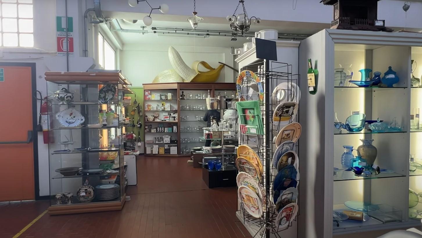 A second-hand or antique store interior with various glass display cases and shelves filled with ceramic plates, glassware, and household items. A large decorative banana sculpture is positioned on top of a cabinet in the background. The store has a warm, vintage atmosphere with wooden flooring and soft lighting.